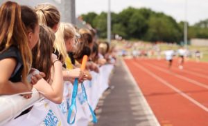 A group of children watching at the side of a track whilst a 100m race is taking place.